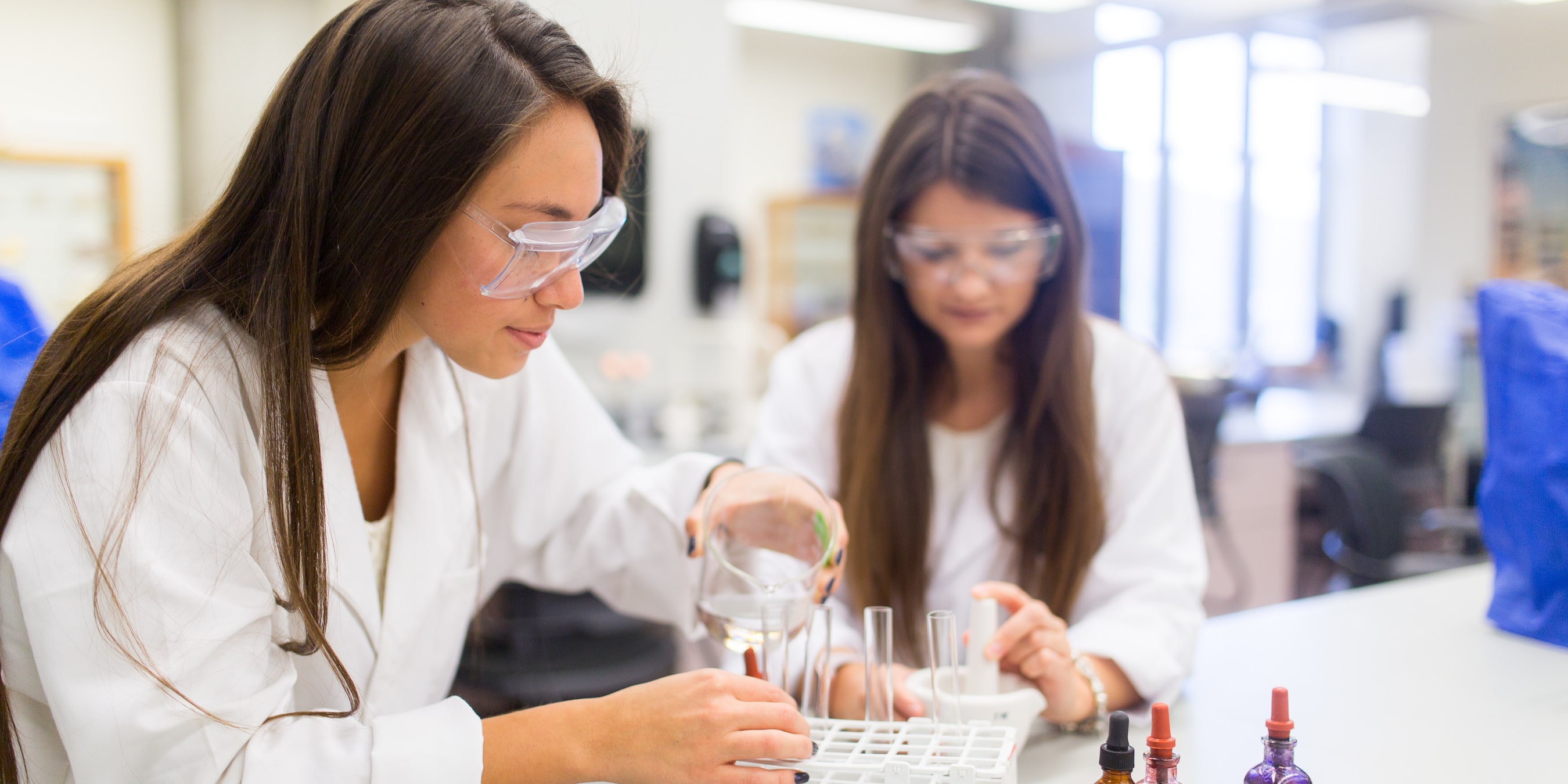 two female students wearing lab coats and safety glasses while pouring liquid in beaker