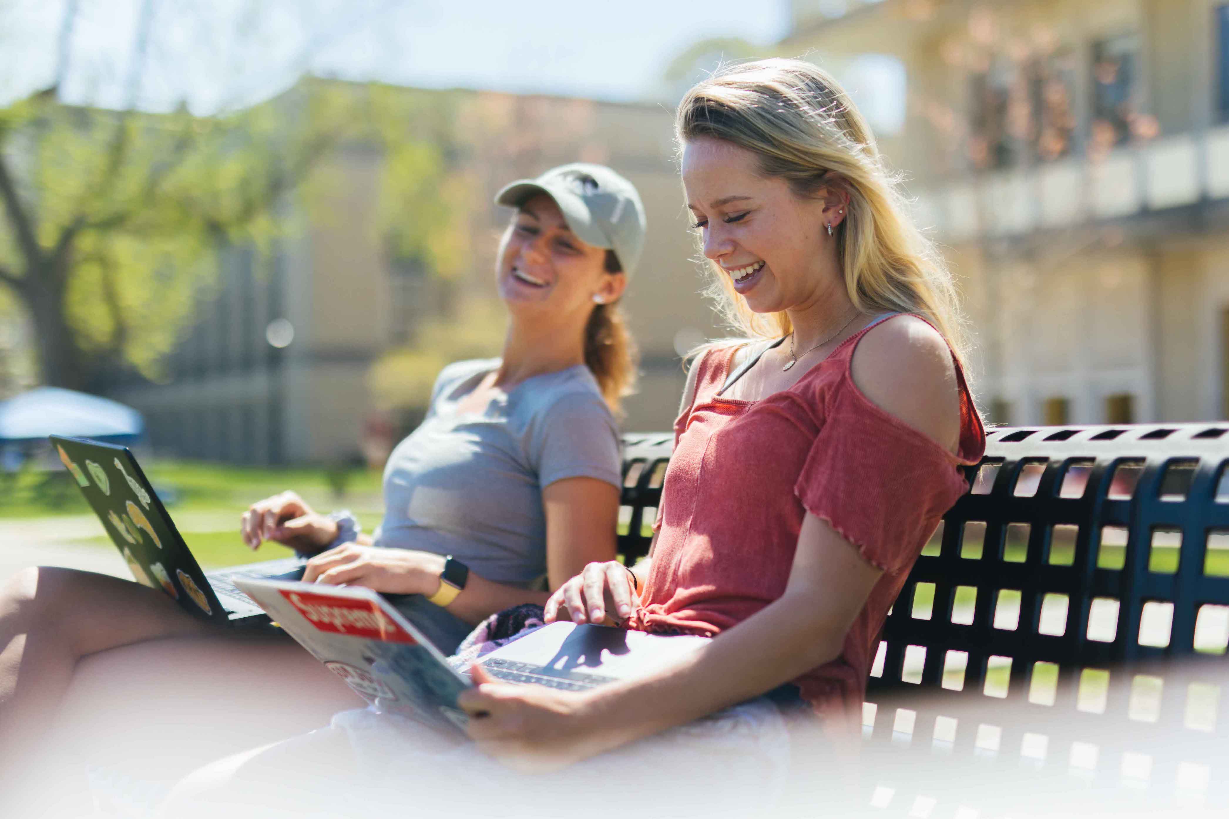 two females laughing outside on a bench while using laptops