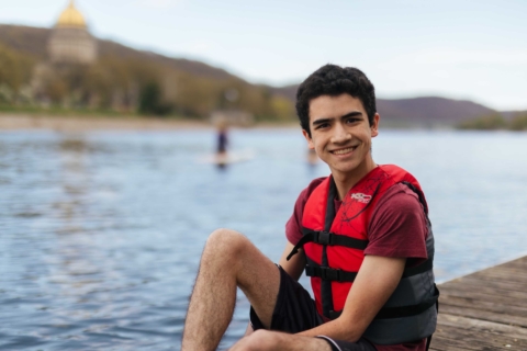 Male student sitting on boat dock at the river wearing a life jacket