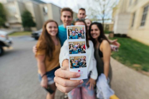 male and female students standing together while female student holds up three photobooth pictures of the group