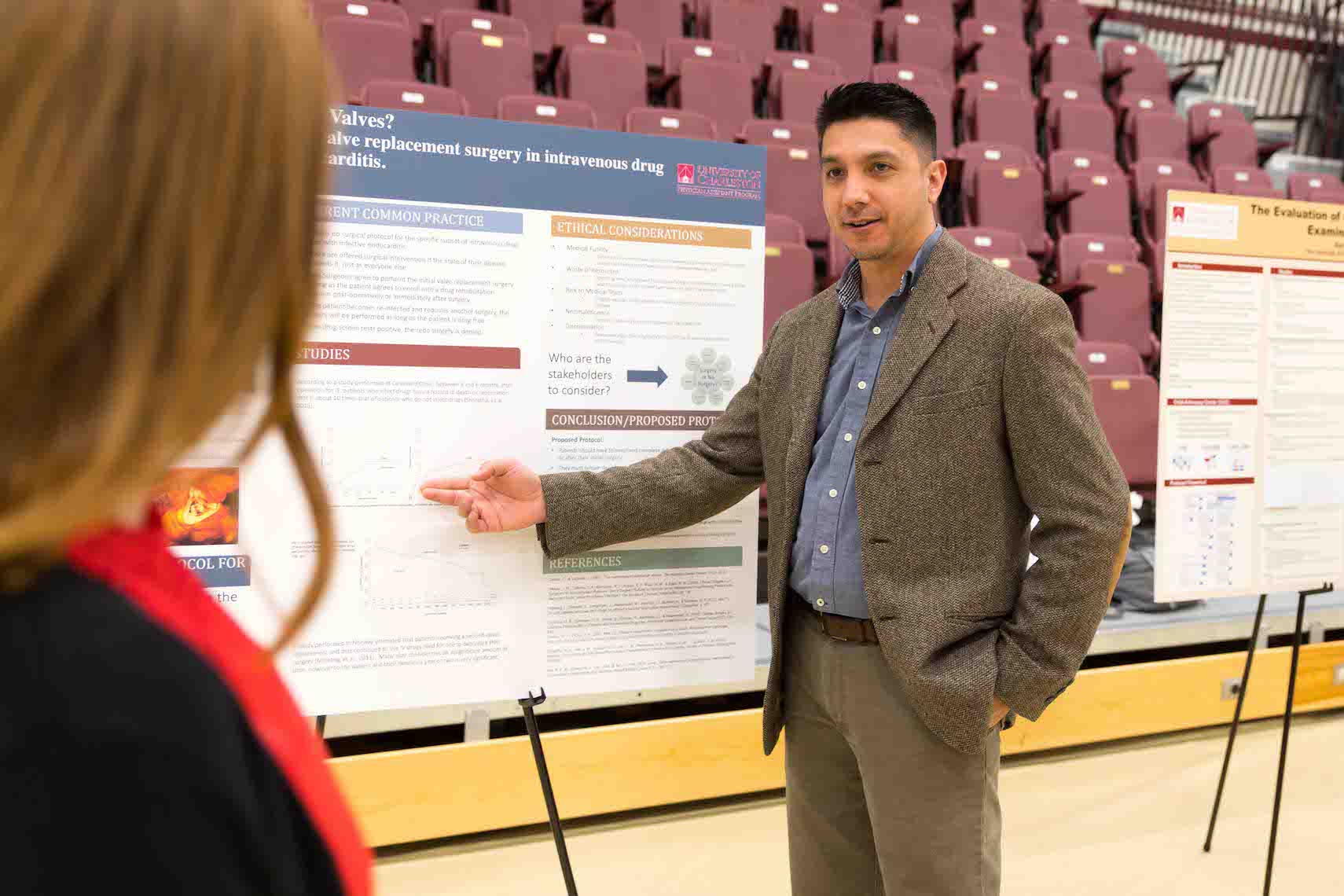 Male student in professional attire pointing to and discussing his research poster