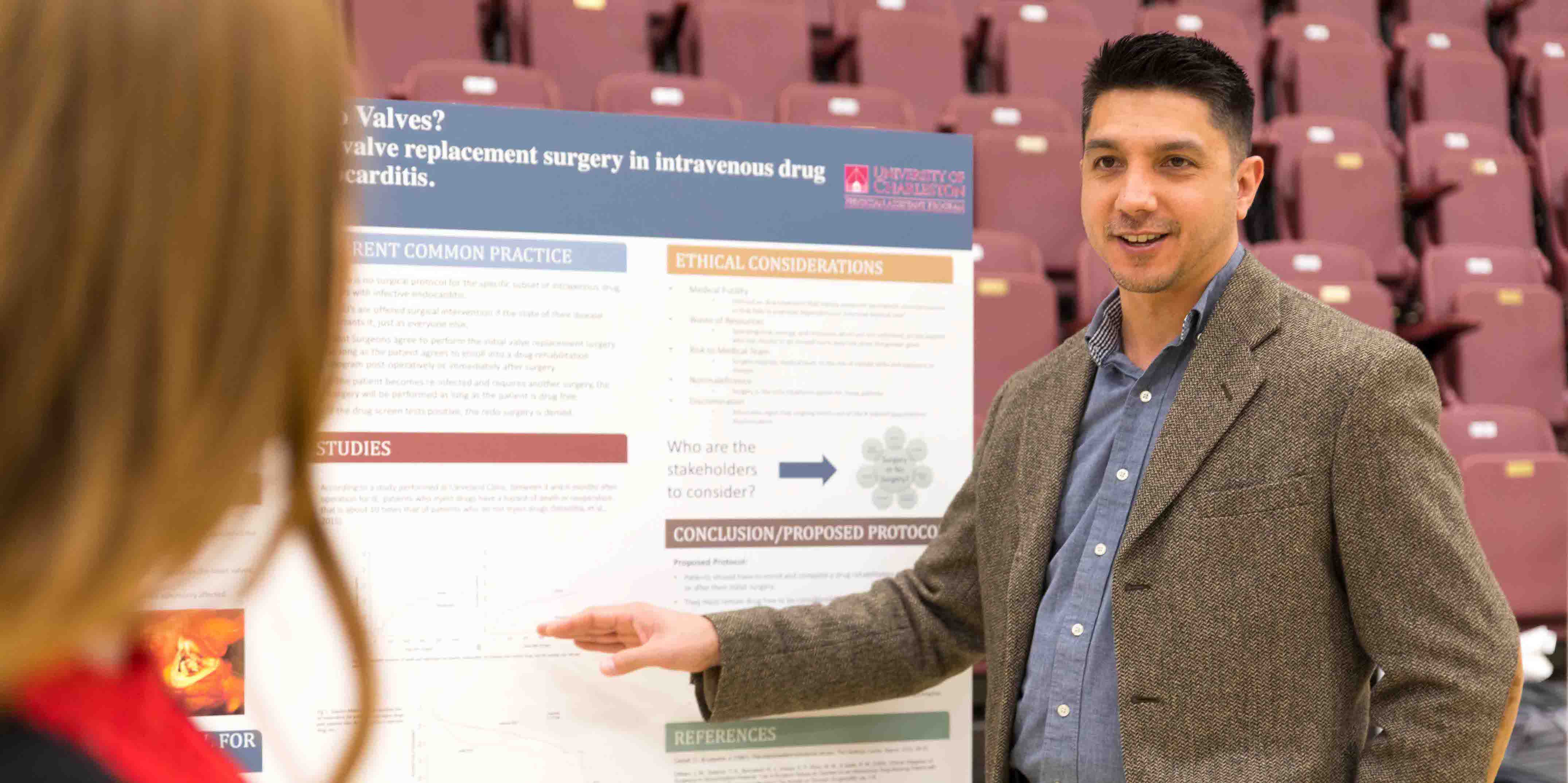 Male student in professional attire pointing to and discussing his research poster