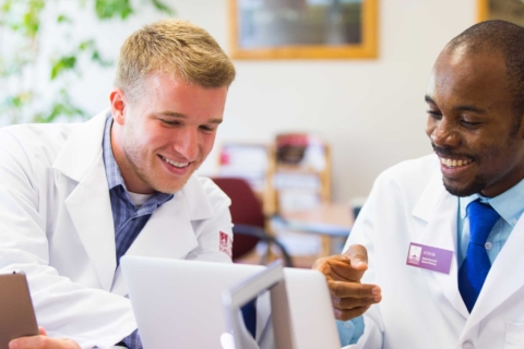 two male pharmacy students in lab coats looking at document on laptop and tablet