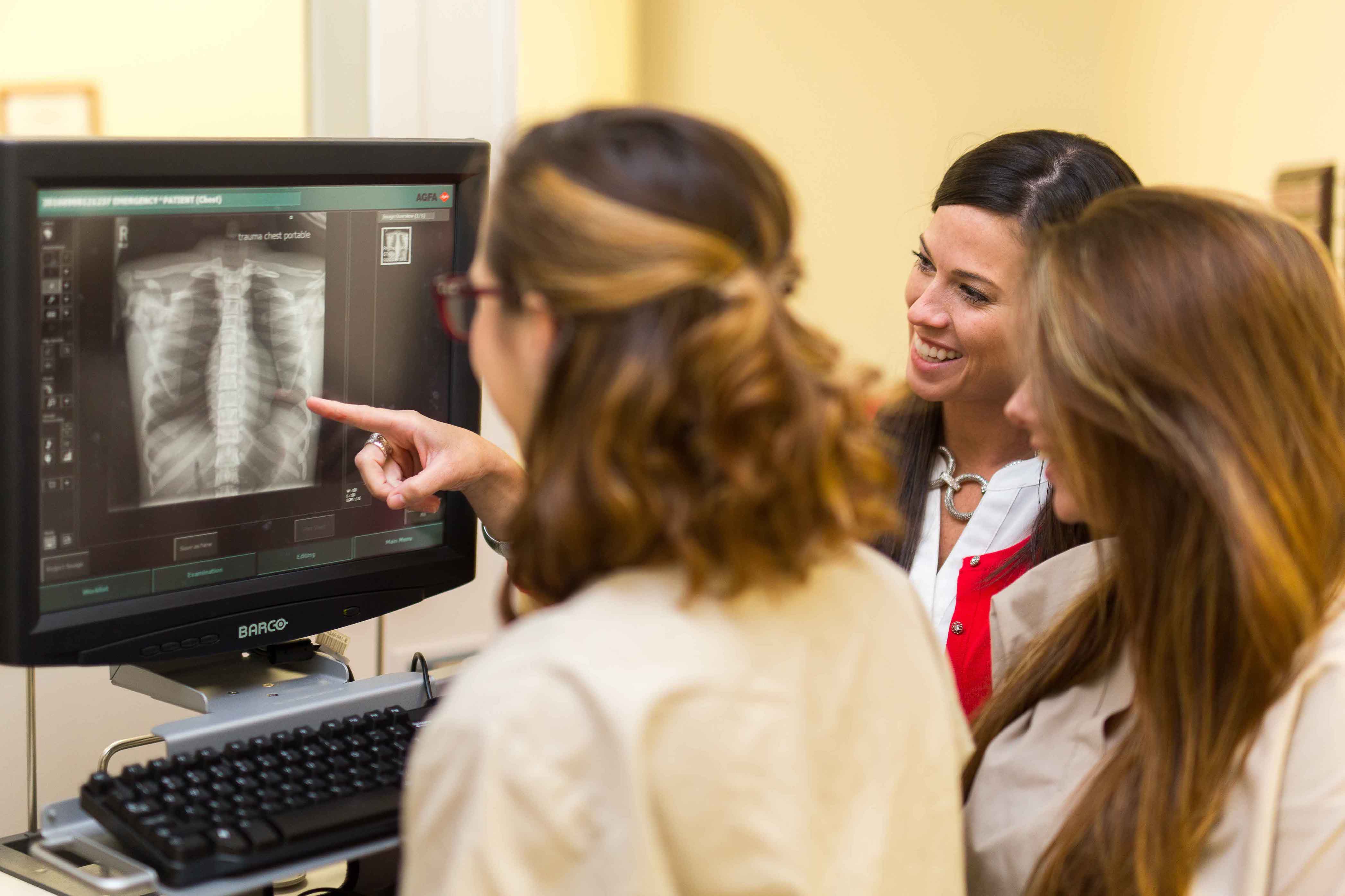 two female radiology students in scrubs looking at x-ray image on computer while professor points to image