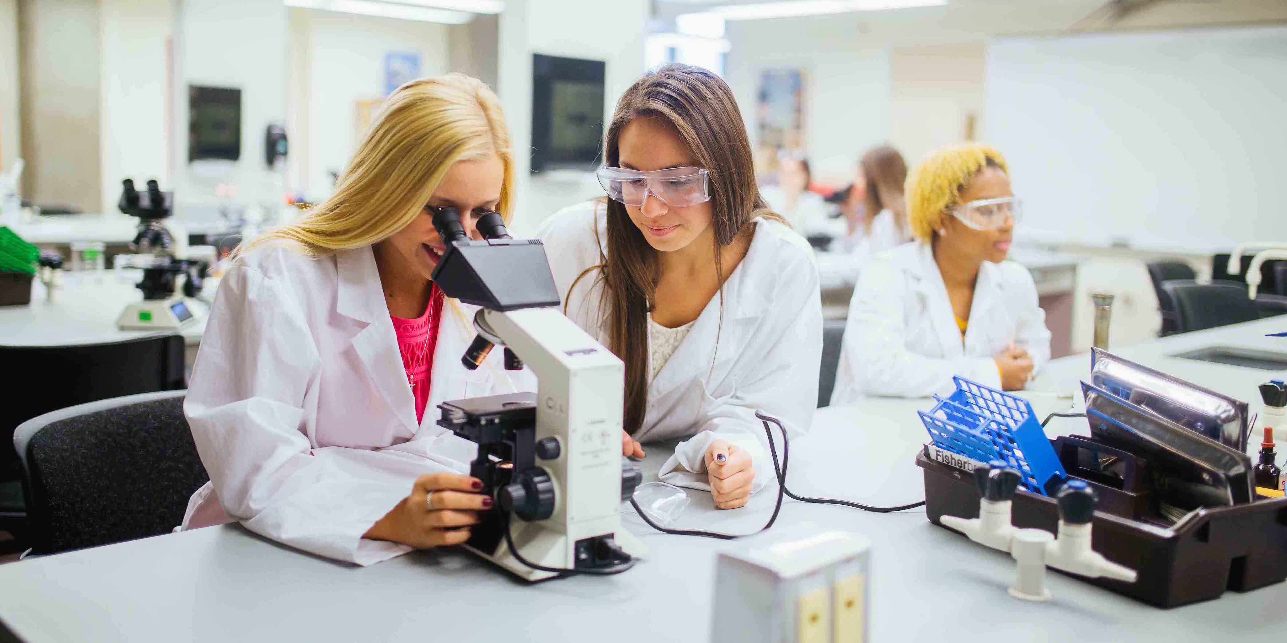 three female students in lab wearing lab coats and safety glasses while using microscope