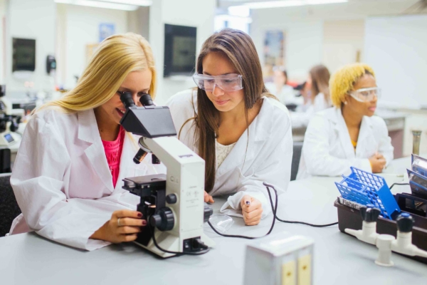 three female students in lab wearing lab coats and safety glasses while using microscope