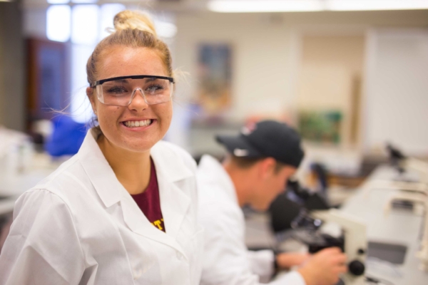 smiling female student in lab wearing lab coat and safety glasses