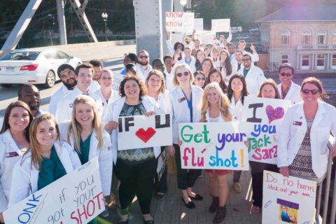 Group of pharmacy students in lab coats standing on bridge with signs to promote getting your flu shot