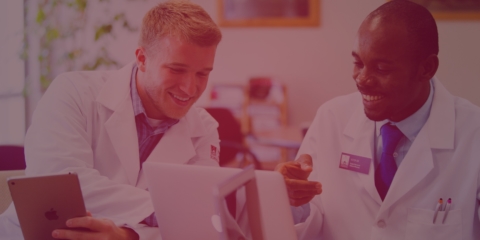 two male pharmacy students in lab coats working on tablet and computer