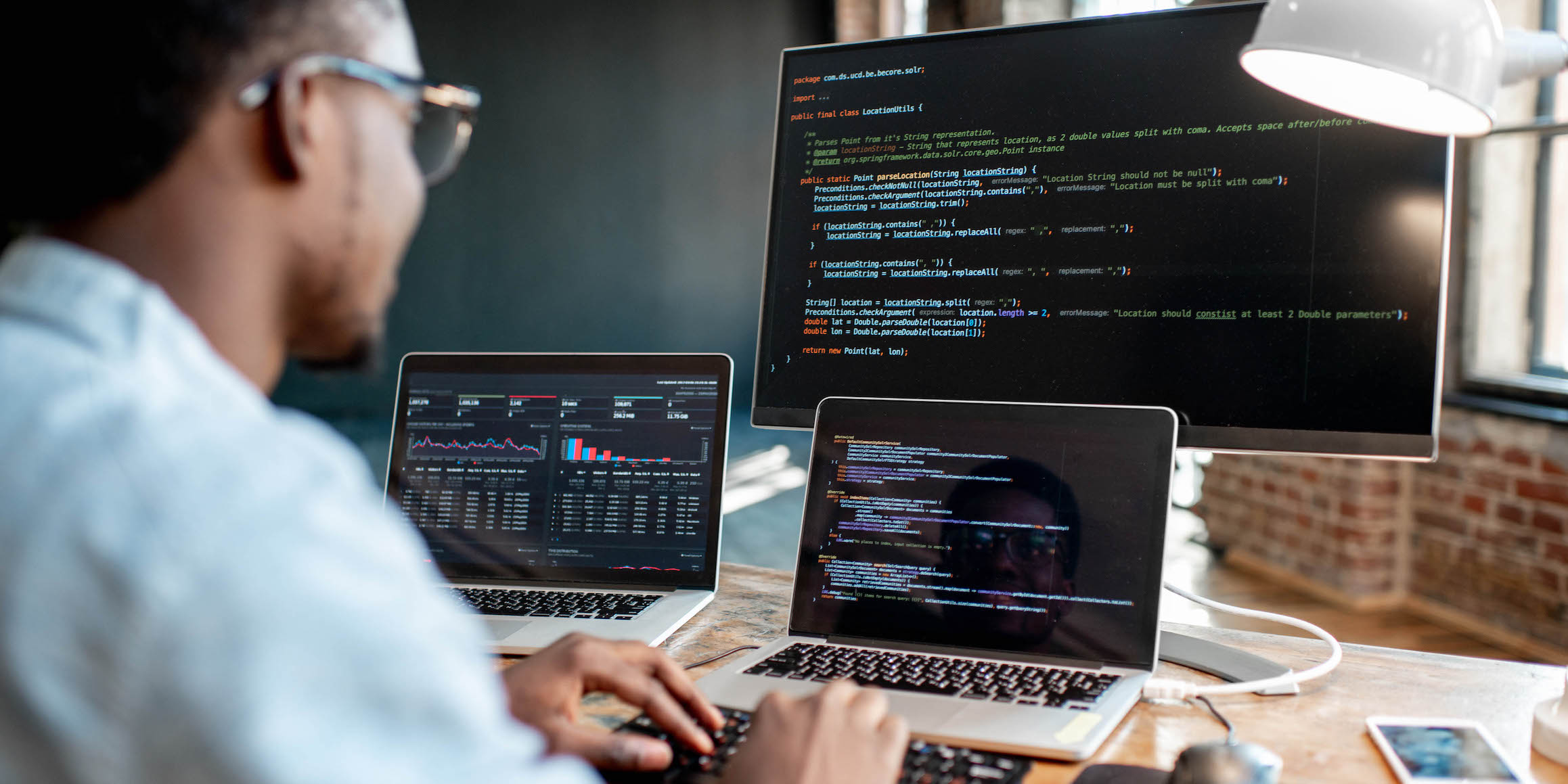 Young african male programmer writing program code sitting at the workplace with three monitors in the office. Image focused on the screen